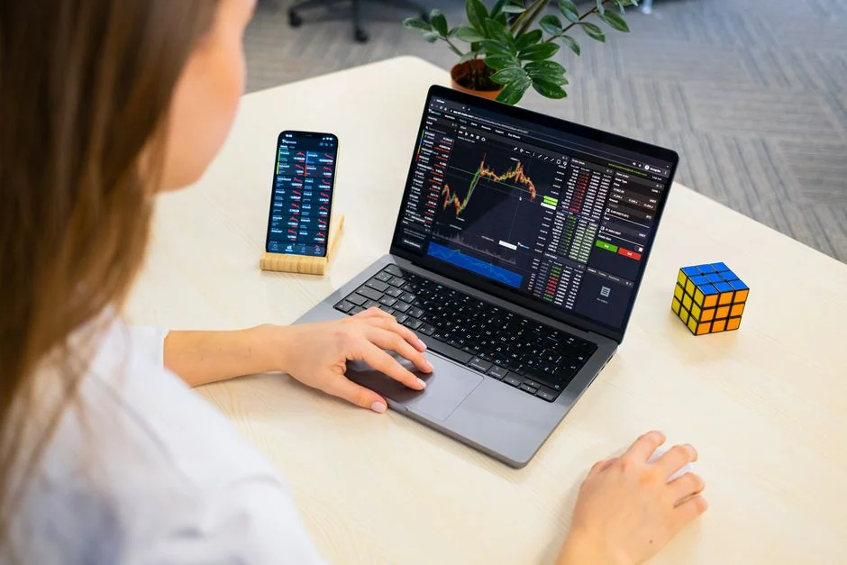 Hands using smartphone beside laptop with stock charts, showcasing digital trading.